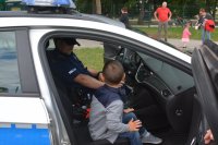 Obraz przedstawia dzielnicowego w radiowozie policyjnym na imprezie plenerowej w Czyżewie pokazującego dzieciom wyposażenie pojazdu