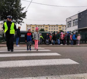 Obraz przedstawia policjanta przeprowadzającego dzieci przez przejście dla pieszych