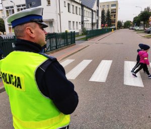 Obraz przedstawia policjanta przeprowadzającego dzieci przez przejście dla pieszych