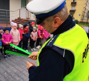 Obraz przedstawia policjanta trzymającego odblaski w tle widać dzieci. Na odblasku napis Urząd Miasta Wysokie Mazowieckie