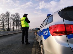 Obraz przedstawia policjanta Wydziału Ruchu Drogowego dokonującego pomiaru prędkości na terenie gminy Wysokie Mazowieckie. W tle widać radiowóz oraz jadące pojazdy