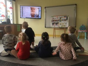 Obraz przedstawia dzieci siedzące na dywanie patrzące w monitor komputera na profilaktyk prowadzącą zajęcia
