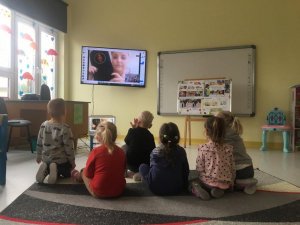 Obraz przedstawia dzieci siedzące na dywanie patrzące w monitor komputera na profilaktyk prowadzącą zajęcia