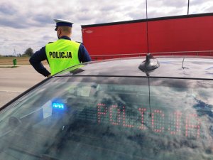 Obraz przedstawia radiowóz policji z wyświetlającym się napisem Policja. W tle widać policjanta i przejeżdżający pojazd