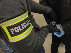 Obraz przedstawia dłonie zatrzymanego w kajdankach i ramię policjanta z opaską Policja