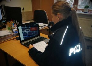 Obraz przedstawia policjantkę siedzącą przed laptopem