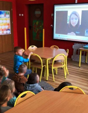 Obraz przedstawia dzieci patrzące w ekran telewizora, gdzie na monitorze widać policjantkę prowadzącą zajęcia online