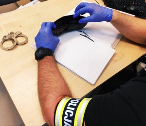 Obraz przedstawia dłonie  policjanta z w rękawiczkach trzymających portfel. W tle  kajdanki i laptop