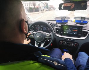 Obraz przedstawia policjanta siedzącego za kierownicą radiowozu