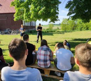 obraz przedstawia policjantkę i profilaktyk na spotkaniu z uczniami we Wnorach Kużelach, w tle dzieci i rowery