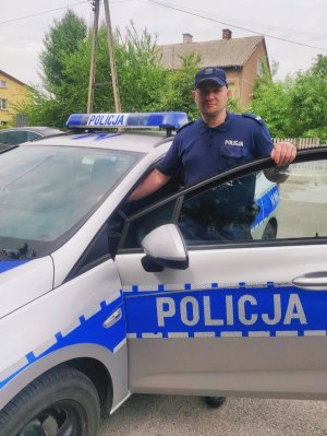 Obraz przedstawia policjanta dzielnicowego Waldemara Włostowskiego stojącego przy radiowozie