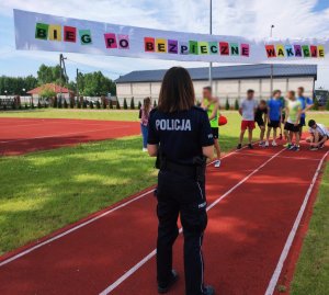 obraz przedstawia policjantkę, w tle uczniowie biorący udział w biegu oraz napis bieg po bezpieczne wakacje