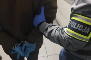 obraz przedstawia ramię mężczyzny, na którym widnieje opaska z napisem policja trzymające nadgarstki mężczyzny  w kajdankach