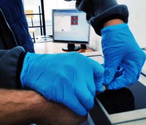 obraz przedstawia dłonie męskie w niebieskich  w rękawiczkach przykładające palec dłoni męskiej do czarnej szklanej płytki, w tle monitor komputera