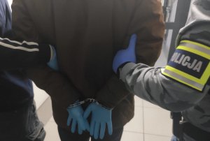 obraz przedstawia ramiona policjantów z opaską z napisem Policja prowadzących zatrzymanego w kajdankach