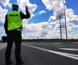 obraz przedstawia policjanta umundurowanego stojącego tyłem trzymającego dłoń w górze, w drugiej ręce trzyma urządzenie do pomiaru prędkości