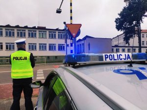 obraz przedstawia policjanta sprawdzającego oznakowanie przy szkole w Wysokiem Mazowieckim, w tle pojazdy i budynek szkoły i radiowóz