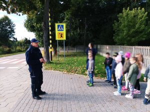 obraz przedstawia policjanta na spotkaniu z dziećmi, w tle znak drogowy