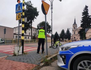 obraz przedstawia policjanta ruchu drogowego stojącego przed przejściem dla pieszych, w tle pojazdy, kościół, rowerzysta