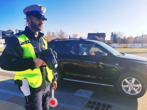 obraz przedstawia policjanta w kamizelce odblaskowej trzymającego w ręce urządzenie do pomiaru prędkości, w tle pojazdy