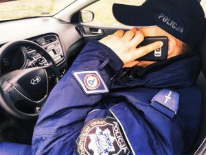 obraz przedstawia policjanta siedzącego w radiowozie trzymającego w ręce telefon