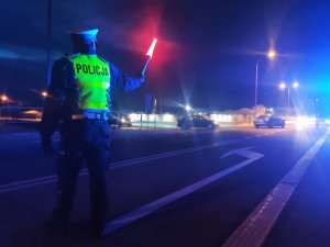 obraz przedstawia policjanta w kamizelce odblaskowej z latarką w ręku, w tle pojazdy