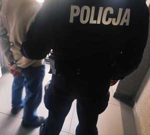 obraz przedstawia policjanta trzymającego za ramię mężczyznę w kajdankach