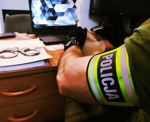 obraz przedstawia policjanta pakującego narkotyki, w tle monitor i kajdanki