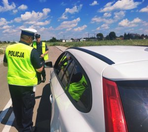 obraz przedstawia policjantów, w tle pojazd koloru białego