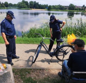 obraz przedstawia policjantów stojących przy rowerze, w tle zbiornik wodny oraz mężczyzna siedzący na ławce