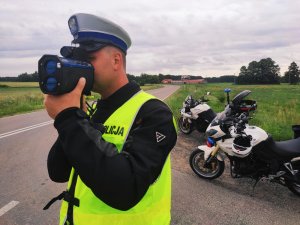 obraz przedstawia policjanta trzymającego w dłoniach urządzenie do pomiaru prędkości, w tle motocykle