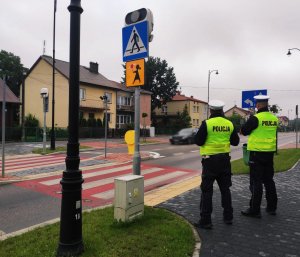 obraz przedstawia policjantów sprawdzających oznakowanie przy szkole w Wysokiem Mazowieckim, w tle pojazdy i budynek szkoły