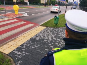 obraz przedstawia policjanta sprawdzającego oznakowanie przy szkole w Wysokiem Mazowieckim, w tle pojazdy i budynek szkoły