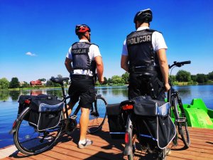 obraz przedstawia policjantów na rowerach na tle zbiornika wodnego w Ciechanowcu