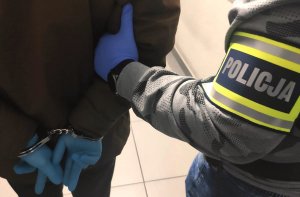 obraz przedstawia ramię męskie z opaską na ramieniu z napisem policja trzymające dłonie męskie w rękawiczkach i kajdankach