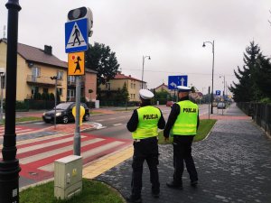 obraz przedstawia policjantów ruchu drogowego stojących przed przejściem dla pieszych, w tle pojazdy