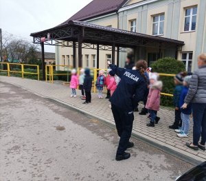 obraz przedstawia policjantkę stojącą przed szkołą pokazującą znak drogowy, w tle dzieci