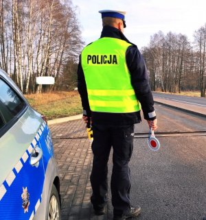 obraz przedstawia policjanta trzymającego w ręce urządzenie do badania stanu trzeźwości i tarcze do zatrzymywania pojazdów, w tle radiowóz