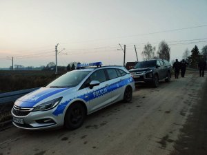 obraz przedstawia policjanta i strażników ochrony kolei na tle przejazdu kolejowego, w tle radiowóz oraz pojazd strażników