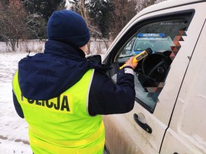 obraz przedstawia policjanta trzymającego w ręce urządzenie do pomiaru stanu trzeźwości, po prawej stronie obrazu pojazd w kolorze białym, w tle radiowóz