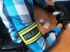 obraz przedstawia ramię mężczyzny, na którym widnieje opaska z napisem policja, oraz dłonie w rękawicach trzymające torebkę foliową z zawartością białego proszku