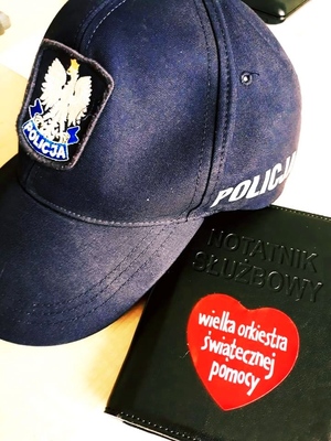 obraz przedstawia czapkę policjanta oraz notatnik służbowy na którym widnieje serduszko Wielkiej Orkiestry Świątecznej Pomocy