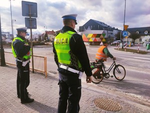 obraz przedstawia policjantów stojących przed przejściem dla pieszych, w tle budynki mieszkalne oraz rowerzysta