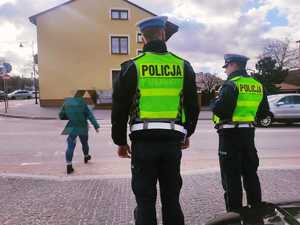 obraz przedstawia policjantów stojących przed przejściem dla pieszych, w tle budynki mieszkalne oraz pieszy