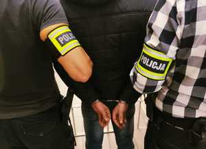 obraz przedstawia dwóch mężczyzn z opaskami na ręce z napisem policja oraz mężczyznę z zapiętymi kajdankami na nadgarstkach