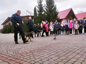 obraz przedstawia policjantów na spotkaniu z dziećmi, policjant przewodnik prowadzi psa służbowego