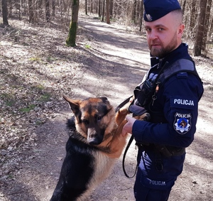 obraz przedstawia policjanta z psem służbowym na tle masywu leśnego