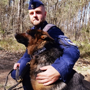 obraz przedstawia policjanta z psem służbowym na tle masywu leśnego