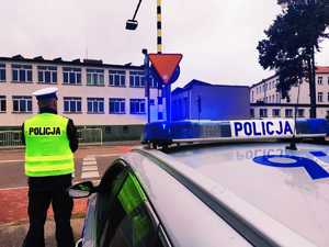 obraz przedstawia policjanta stojącego przed przejściem dla pieszych,  w tle budynek szkoły i radiowóz