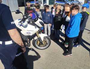 obraz przedstawia policjanta z dziećmi na placu komendy, w tle motocykl